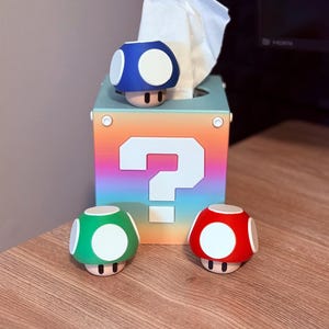 May include: A rainbow-colored question mark box with a white question mark on the front. The box is a tissue box with a blue, green, and red mushroom on top.