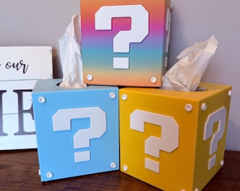 May include: Three tissue box covers shaped like question mark blocks from the Super Mario Bros. video game. The top box is a rainbow gradient, the middle box is light blue, and the bottom box is yellow.