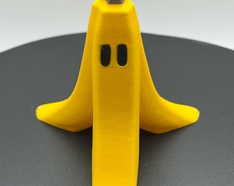May include: A 3D-printed yellow banana peel with two black oval eyes and a brown stem. The peel is open, revealing a cartoon-like design. The object is on a dark gray surface.