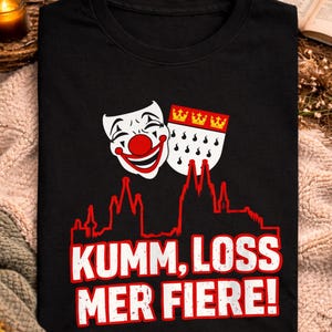 Puede incluir: Camiseta negra con un diseño que incluye una máscara de payaso, un escudo de armas y el texto "Kumm, Loss Mer Fiere!" en blanco. La palabra "schwarz" está en la parte superior. La camiseta se muestra sobre una superficie texturizada.