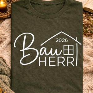 Peut inclure: T-shirt vert foncé avec l'inscription blanche "Bauherr 2026" et un dessin de maison. Le t-shirt est présenté sur une couverture, avec une tasse de café, une bougie et un livre ouvert en arrière-plan.
