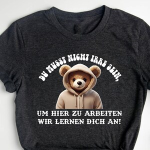 Può includere: T-shirt grigio scuro con un orso dei cartoni animati con una felpa con cappuccio. La maglietta ha un testo bianco che recita "DU MUSST NICHT IRRE SEIN, UM HIER ZU ARBEITEN WIR LERNEN DICH AN!" Le parole "dunkles heidekraut" sono in alto.