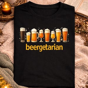 Peut inclure: T-shirt noir avec un motif de différents verres à bière avec de la mousse. Le mot "beergetarian" est imprimé en jaune sous les verres à bière. Le t-shirt est présenté sur une surface texturée avec une tasse, une bougie et un livre en arrière-plan.