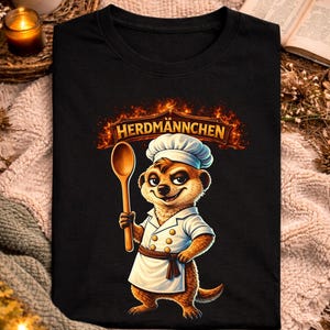 Op de afbeelding: Zwart T-shirt met een cartoon stokstaartje chef-kok met een houten lepel. Het shirt heeft de tekst "HERDMANNCHEN" boven het stokstaartje. Het woord "schwarz" staat bovenaan het shirt.