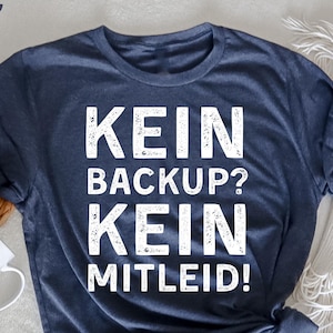 Può includere: Una maglietta blu navy con la scritta bianca "KEIN BACKUP? KEIN MITLEID!" in un carattere invecchiato. La maglietta è annodata sull'orlo. Una tazza bianca e un sottobicchiere intrecciato sono visibili sullo sfondo.