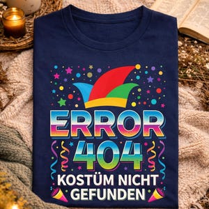 May include: A navy blue t-shirt featuring the text "ERROR 404 KOSTÜM NICHT GEFUNDEN" in a colourful design with a jester's hat, stars, confetti, and streamers. The shirt is displayed on a neutral-coloured blanket.