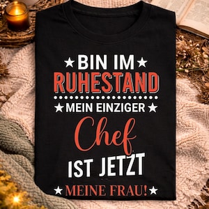 Peut inclure: T-shirt noir avec texte allemand "BIN IM RUHESTAND MEIN EINZIGER Chef IST JETZT MEINE FRAU!" en blanc et rouge. Le t-shirt est présenté sur une surface texturée avec une tasse de café, une bougie et un livre ouvert.