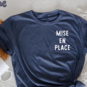 Peut inclure: Un t-shirt bleu marine avec le texte "MISE EN PLACE" en blanc. Le mot "marine" est en haut à gauche. Le t-shirt est stylisé avec une manche roulée et un nœud en bas. Une tasse blanche et un tapis tissé sont en arrière-plan.