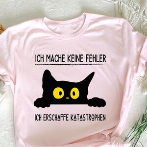 May include: A pale pink t-shirt featuring the German text "ICH MACHE KEINE FEHLER" and "ICH ERSCHAFFE KATASTROPHEN", with a black cat graphic with yellow eyes. The t-shirt is folded and knotted at the bottom.