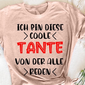 Può includere: T-shirt rosa chiaro con il testo tedesco "ICH BIN DIESE > COOLE < TANTE VON DER ALLE > REDEN <" in nero e rosso. La maglietta è annodata in basso. Una tazza bianca e fiori sono sullo sfondo.
