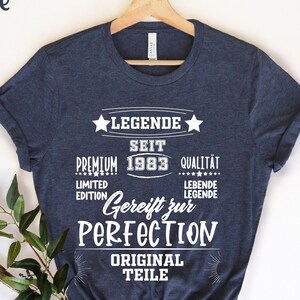 May include: Heather marine navy blue t-shirt with white text. The text includes "LEGENDE SEIT PREMIUM 1983 QUALITAT LIMITED EDITION Gereift zur PERFECTION ORIGINAL TEILE". The shirt is hanging on a wooden hanger.