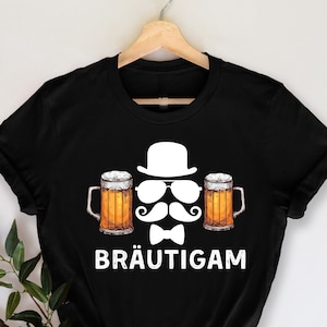 K&ouml;nnte beinhalten: Schwarzes T-Shirt mit einem grafischen Design, das einen wei&szlig;en Hut, eine Sonnenbrille, einen Schnurrbart und eine Fliege zeigt, flankiert von zwei Bierkr&uuml;gen. Das Wort "BR&Auml;UTIGAM" ist darunter gedruckt.