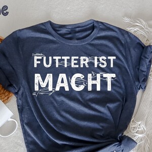 May include: A navy blue t-shirt with the German phrase "Futter ist Macht" in white block letters. The shirt has a crew neck and short sleeves. The word "marine" is in the upper left corner. A white coffee mug and a woven basket are in the background.