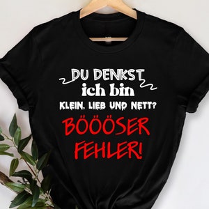 May include: Black t-shirt with the German text "DU DENKST ich bin KLEIN, LIEB UND NETT? BÖÖÖSER FEHLER!" in white and red. The shirt is hanging on a wooden hanger.