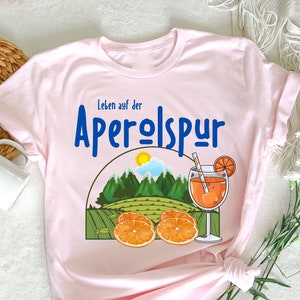May include: Pink t-shirt with a graphic of a glass of Aperol Spritz, oranges, and a mountain scene. The text "Leben auf der Aperolspur" is printed on the shirt.