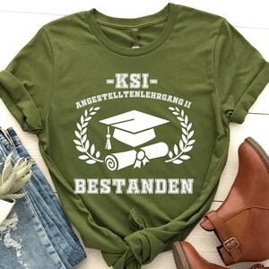 Peut inclure: Un t-shirt vert olive avec du texte et des graphiques blancs, dont une toque de diplômé et un diplôme, et le mot "BESTANDEN". Le t-shirt est associé à un jean bleu et des bottines marron, pour un look décontracté.
