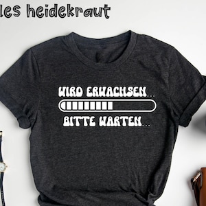 Peut inclure: T-shirt gris foncé avec texte blanc "Wird erwachsen... Bitte warten..." au-dessus d'une barre de chargement. Le t-shirt a des manches retroussées et un nœud en bas. Une montre, des lunettes et une tasse de café sont visibles.
