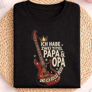 Può includere: T-shirt nero con grafica di chitarra elettrica rossa e marrone e la scritta "ICH HABE ZWEI TITEL PAPA & OPA UND ICH ROCKE BEIDE". La parola "schwarz" è in alto. La maglietta è su un tappetino intrecciato.