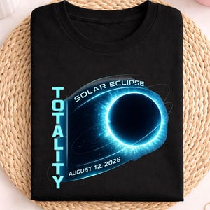 Pode incluir: Camiseta preta com um design de eclipse solar azul e preto. O texto inclui "TOTALITY", "SOLAR ECLIPSE" e "AUGUST 12, 2026". A palavra "schwarz" é impressa na parte superior da imagem.