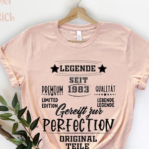 May include: A light pink t-shirt with the text "LEGENDE SEIT 1983 GEREIFT ZUR PERFECTION" in black. The shirt is hanging on a wooden hanger. The text also includes the words "PREMIUM", "QUALIT&Auml;T", "LIMITED EDITION", "LEBENDE LEGENDE", and "ORIGINAL TEILE".