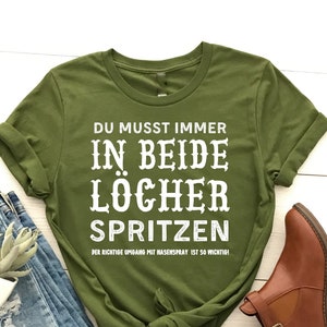 Può includere: Una t-shirt verde oliva con il testo tedesco "DU MUSST IMMER IN BEIDE LÖCHER SPRITZEN" in bianco. La maglietta è abbinata a jeans blu, una piccola pianta e stivaletti marroni.