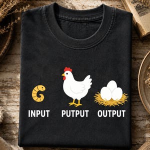 May include: A black t-shirt featuring a graphic print of a yellow worm labeled INPUT, a white chicken labeled PUTPUT, and three white eggs in a nest labeled OUTPUT. The design uses white text and colorful illustrations to show a humorous life cycle.