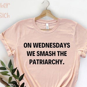 Könnte beinhalten: Hellrosa T-Shirt mit dem schwarzen Text "ON WEDNESDAYS WE SMASH THE PATRIARCHY". Das T-Shirt hängt an einem Holzkleiderbügel. Der Markenname "Heather Pfirsich" ist oben links zu sehen.