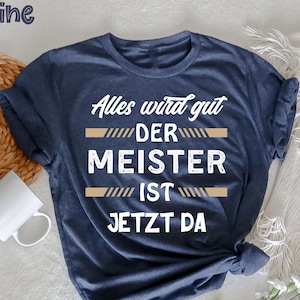 May include: A navy blue t-shirt with the text "Alles wird gut DER MEISTER IST JETZT DA" in white. The text is separated by horizontal tan lines. The word "marine" is in the upper left corner. A white mug and woven mat are in the background.