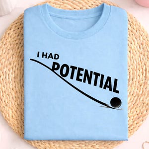 Könnte beinhalten: Hellblaues T-Shirt mit dem schwarzen Text "I HAD POTENTIAL". Das Design beinhaltet eine schwarze Linie und eine schwarze Kugel. Das T-Shirt wird auf einer gewebten Matte präsentiert.
