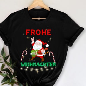 Può includere: T-shirt nera con un design natalizio. Presenta Babbo Natale che suona una chitarra verde, con le parole "FROHE WEIHNACHTEN" in rosso e verde. Bastoncini di zucchero e luci di stringa completano il tema.