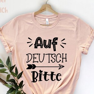 May include: A peach colored t-shirt with black text that reads "Auf Deutsch Bitte" with an arrow pointing to the right.
