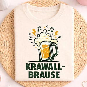 May include: Cream-colored t-shirt featuring a beer stein graphic with musical notes and lightning bolts. The text "KRAWALL-BRAUSE" is printed in green below the graphic. The shirt is displayed on a woven placemat with floral and decorative elements.