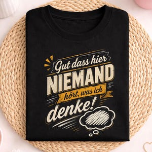 Op de afbeelding: Zwart t-shirt met de Duitse tekst "Gut dass hier NIEMAND hört, was ich denke!" in een verweerde lettertype. Het ontwerp bevat een tekstballon en decoratieve elementen. Het shirt wordt gepresenteerd op een geweven mat.