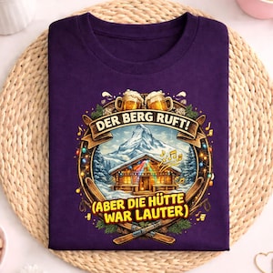 Puede incluir: Una camiseta morada con un diseño gráfico. El diseño incluye el texto "DER BERG RUFT!" y "(ABER DIE HÜTTE WAR LAUTER)", una escena de montaña, jarras de cerveza, esquís y notas musicales. La camiseta se muestra sobre una estera tejida.