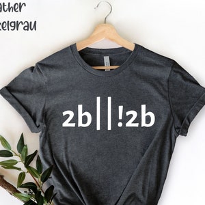 Pode incluir: T-shirt cinza escuro com o texto branco "2b||!2b". As palavras "Heather Dunkelgrau" estão no canto superior esquerdo. A camiseta é exibida em um cabide de madeira contra um fundo branco. Há vegetação no canto inferior esquerdo.