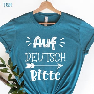 Puede incluir: Una camiseta azul turquesa con el texto "Auf Deutsch Bitte" en letras blancas con una flecha apuntando hacia la derecha. La camiseta está en una percha.