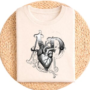 Peut inclure: T-shirt crème avec un motif noir et blanc d'un cœur humain dans les lettres "A" et "P". Le design est orné et de style vintage. Le texte "heather pfirsich" est imprimé en haut.