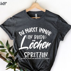 Peut inclure: T-shirt gris foncé chiné avec texte blanc : "DU MUSST IMMER IN BEIDE LÖCHER SPRITZEN". Le texte "DER RICHTIGE UMGANG MIT NASENSPRAY IST SO WICHTIG!" est en dessous. Le t-shirt est suspendu à un cintre en bois.