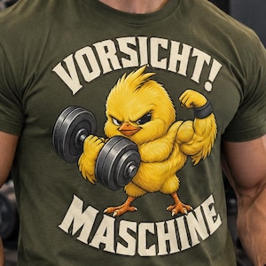 Könnte beinhalten: Ein olivgrünes Kurzarm-T-Shirt mit einer Grafik eines muskulösen gelben Kükens, das eine graue Hantel hebt. Der Text "VORSICHT!" steht über dem Küken und "MASCHINE" darunter in einer fetten, cremeweißen, serifenlosen Schriftart.