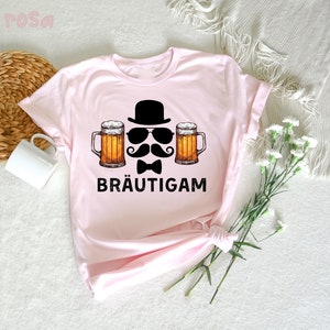 K&ouml;nnte beinhalten: Hellrosa T-Shirt mit einem Aufdruck, der einen schwarzen Zylinder, Sonnenbrille, Schnurrbart, Fliege und zwei Bierkr&uuml;ge zeigt. Das Wort "BR&Auml;UTIGAM" ist in Schwarz gedruckt. Das T-Shirt ist aus weichem Material.