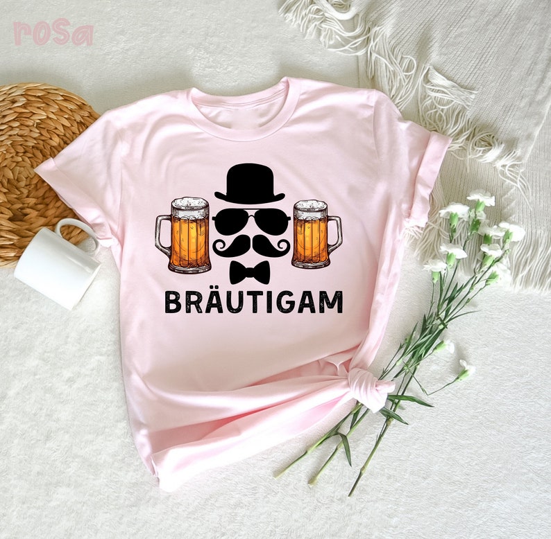 K&ouml;nnte beinhalten: Hellrosa T-Shirt mit einem Aufdruck, der einen schwarzen Zylinder, Sonnenbrille, Schnurrbart, Fliege und zwei Bierkr&uuml;ge zeigt. Das Wort "BR&Auml;UTIGAM" ist in Schwarz gedruckt. Das T-Shirt ist aus weichem Material.