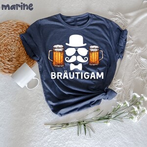 K&ouml;nnte beinhalten: Marineblaues T-Shirt mit einer Grafik von Hut, Sonnenbrille, Schnurrbart, Fliege und zwei Bierkr&uuml;gen. Das Wort "BR&Auml;UTIGAM" ist unter der Grafik aufgedruckt. Das Wort "marine" befindet sich in der oberen linken Ecke.
