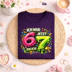 Peut inclure: Un t-shirt violet avec l'inscription allemande "ICH WAR 6 JETZT 7 BIN ICH" dans un style color&eacute; et enfantin. Les chiffres 6 et 7 sont en rose et vert, avec des &eacute;claboussures.
