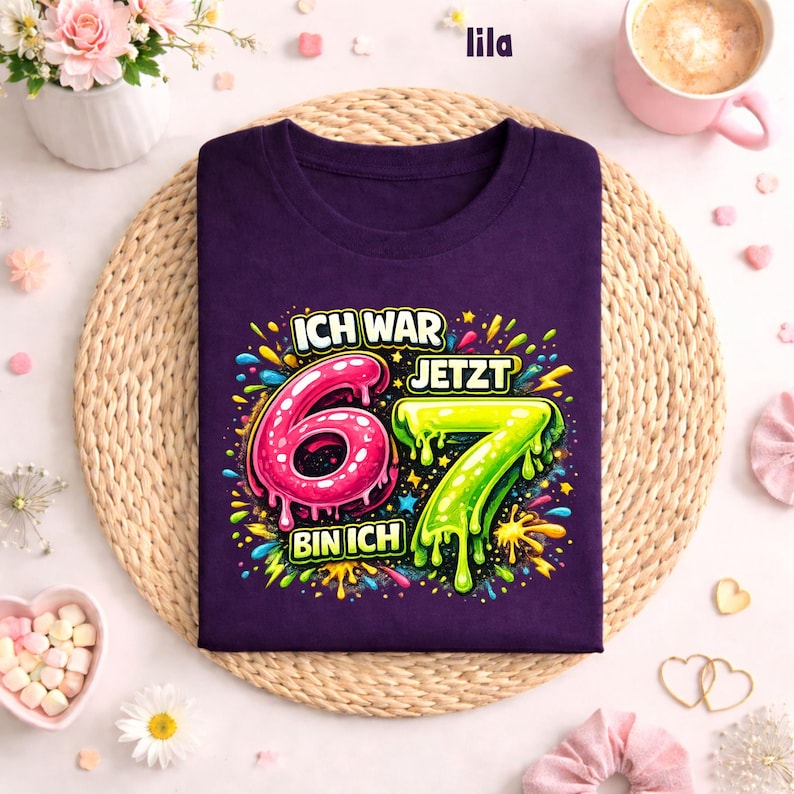 Peut inclure: Un t-shirt violet avec l'inscription allemande "ICH WAR 6 JETZT 7 BIN ICH" dans un style color&eacute; et enfantin. Les chiffres 6 et 7 sont en rose et vert, avec des &eacute;claboussures.