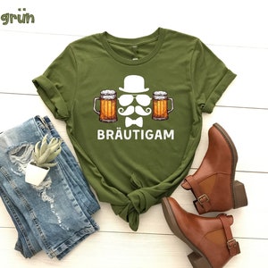 K&ouml;nnte beinhalten: Olivgr&uuml;nes T-Shirt mit einer Grafik von Hut, Sonnenbrille, Schnurrbart, Fliege und zwei Bierkr&uuml;gen. Das Wort "BR&Auml;UTIGAM" ist darunter gedruckt. Das Shirt ist mit blauen Jeans und braunen Stiefeletten gestylt.