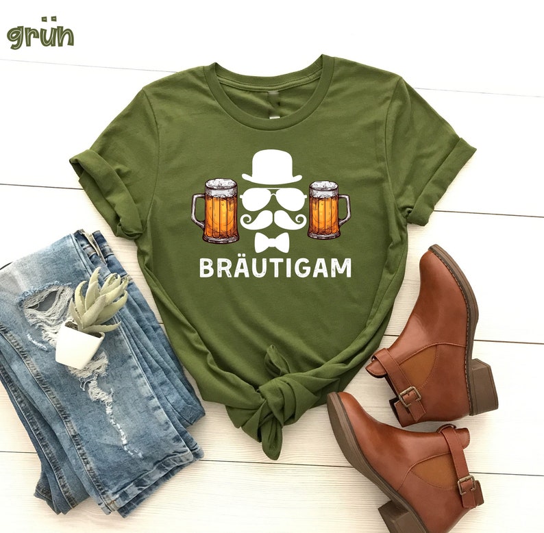 K&ouml;nnte beinhalten: Olivgr&uuml;nes T-Shirt mit einer Grafik von Hut, Sonnenbrille, Schnurrbart, Fliege und zwei Bierkr&uuml;gen. Das Wort "BR&Auml;UTIGAM" ist darunter gedruckt. Das Shirt ist mit blauen Jeans und braunen Stiefeletten gestylt.