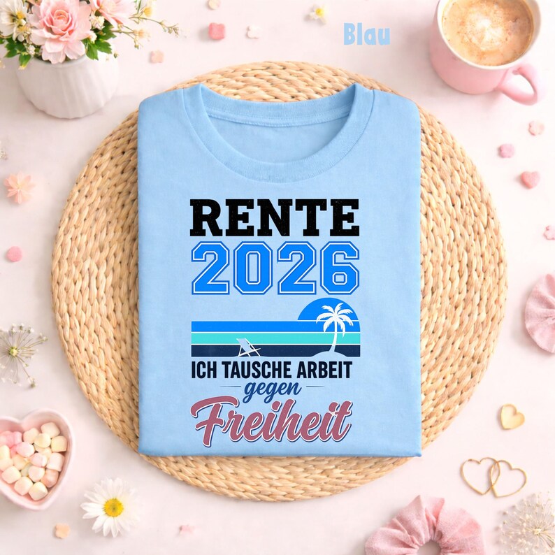 May include: A light blue t-shirt with the words "RENTE 2026" in black and blue. Below the text is a graphic of a beach scene with a palm tree and the words "ICH TAUSCHE ARBEIT gegen Freiheit" in German. The shirt is on a woven mat.