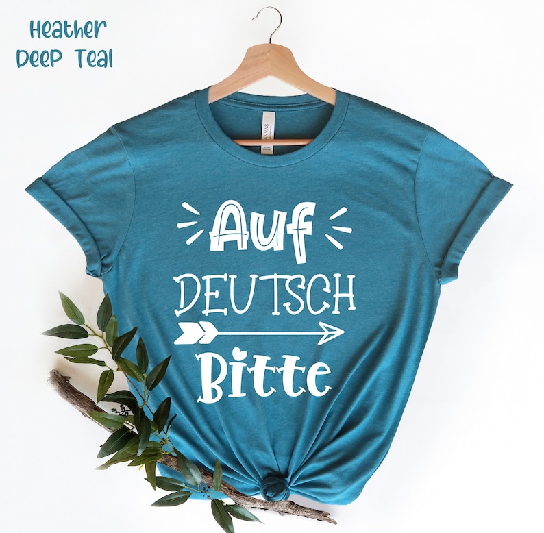 May include: A teal blue t-shirt with the text "Auf Deutsch Bitte" in white lettering with an arrow pointing to the right. The shirt is on a hanger.