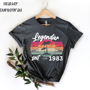May include: Dark gray t-shirt with a retro sunset design featuring palm trees and the text "Legend&auml;r seit 1983". The shirt is hanging on a wooden hanger. The words "Heather Dunkelgrau" are at the top left.