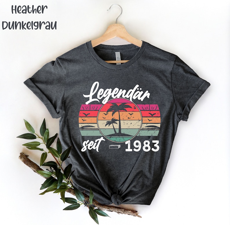 May include: Dark gray t-shirt with a retro sunset design featuring palm trees and the text "Legend&auml;r seit 1983". The shirt is hanging on a wooden hanger. The words "Heather Dunkelgrau" are at the top left.